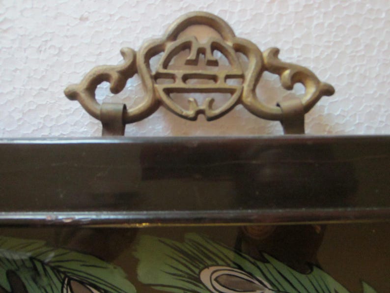 May include: A dark brown lacquered tray featuring a decorative brass handle with an intricate openwork design. The handle is attached to the top edge of the rectangular tray, which displays a partial green and black peacock feather pattern.