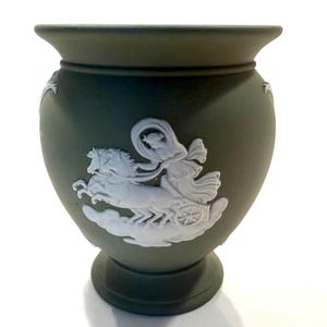 Puede incluir: Un jarrón verde con un diseño en bajorrelieve blanco de un carro tirado por caballos. El jarrón tiene un cuerpo redondeado, un borde acampanado y una base de pedestal. El bajorrelieve es de diseño clásico.