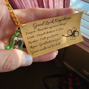 May include: A brown paper tag with the text "Good Luck Keychain" and a list of herbs and their properties. The tag is attached to a small glass jar filled with dried herbs and a green four-leaf clover charm.