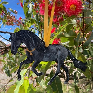 May include: Black horse ornament hanging from a branch of a tree with red flowers. The ornament is made of wood and has a gold ribbon.