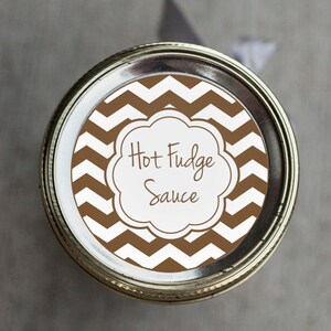 Pode incluir: Uma tampa de frasco com um padrão de chevron marrom e branco com um rótulo branco que diz "Hot Fudge Sauce" em escrita cursiva marrom.