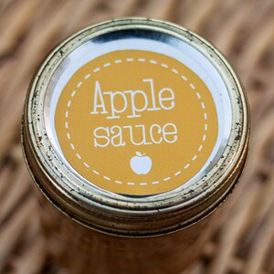 Op de afbeelding: Een glazen pot met een gouden deksel en een geel etiket waarop "Apple sauce" staat met een klein appelpictogram. De pot staat op een geweven rieten oppervlak.
