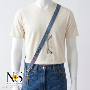 May include: A blue denim lanyard worn across the torso, featuring the embroidered text Mr. Vince and a small red apple icon. The strap includes a metal clip holding a set of silver keys and a pair of thin-rimmed glasses hanging from the fabric.