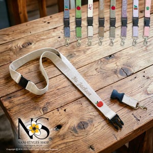Breakaway Safety lanyard with an embroidered personalized name and an embroidered red apple icon. The lanyard has a black plastic clasp and a metal hook. Several other lanyards with different colors and designs are visible in the background.