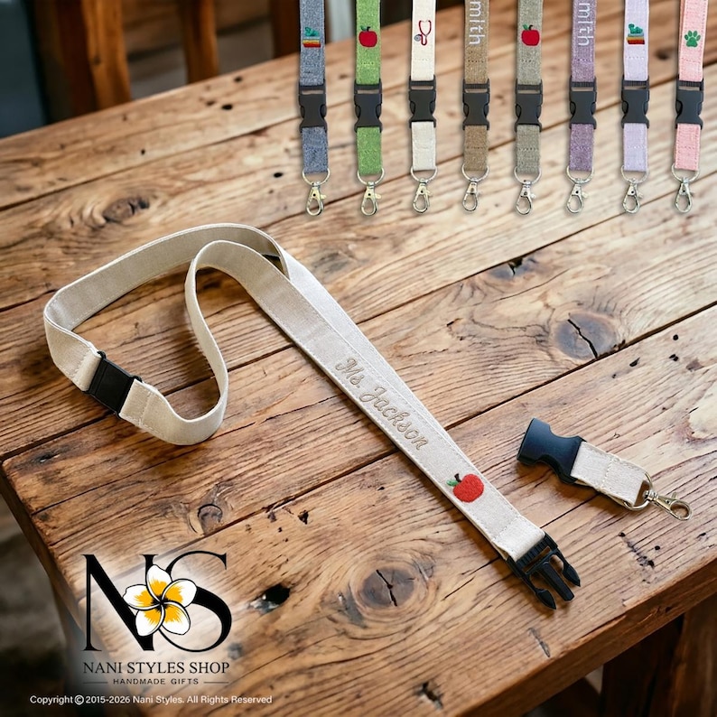 Breakaway Safety lanyard with an embroidered personalized name and an embroidered red apple icon. The lanyard has a black plastic clasp and a metal hook. Several other lanyards with different colors and designs are visible in the background.