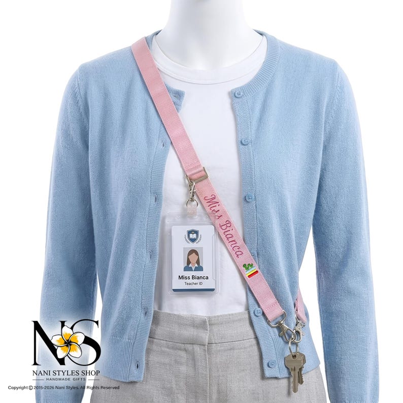 May include: A light pink fabric lanyard featuring the embroidered name Miss Bianca and a small green and red graphic. The lanyard is attached to a clear plastic ID badge holder containing a teacher identification card and a silver metal key ring.