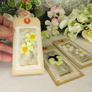 Puede incluir: Etiquetas de regalo hechas a mano con dise&ntilde;os florales. Las etiquetas son rectangulares con un fondo beige y un borde de tela blanca. Cada etiqueta presenta una flor diferente: narcisos, margaritas y pensamientos. Las flores son de fieltro y tienen acentos amarillos y morados.