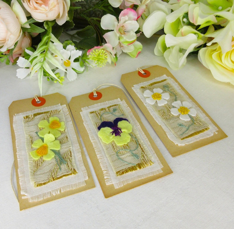 Puede incluir: Tres etiquetas de regalo hechas a mano con dise&ntilde;os florales. Cada etiqueta es rectangular, hecha de cartulina marr&oacute;n, y presenta un panel de tela con flores bordadas. Las flores incluyen narcisos amarillos, un pensamiento morado y amarillo, y margaritas blancas.