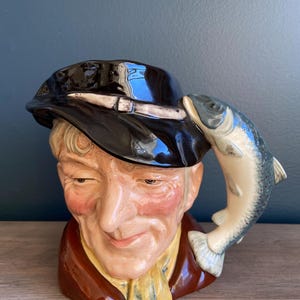 Puede incluir: Un jarro de cerámica con forma de cabeza de hombre que lleva un sombrero negro y sostiene un pez en la mano. El hombre tiene barba y bigote marrones y sonríe. El pez es de color azul claro y gris.