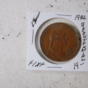 May include: A copper-colored coin from 1932, encased in a white holder. The coin features a profile of a person and the year 1932. The holder has handwritten text, including "Bicentennial" and "FIXF".