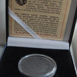 May include: A circular silver coin encased in a clear protective capsule, housed within a black velvet-lined display box. The interior lid features a printed card titled The Imperial Japanese Silver Coin - 50 Sen with descriptive historical text.