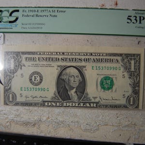 May include: A one-dollar Federal Reserve Note, featuring George Washington's portrait, is encased in a protective sleeve. The note has the serial number E15370990G and is labeled as a Fr. 1910-E 1977A S1 Error.