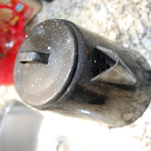 May include: A black enamel coffee pot with a lid and handle. The pot has a cylindrical shape and a spout. The surface has white speckles. The pot is sitting on a speckled countertop.