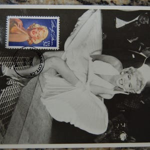May include: Black and white postcard featuring a photograph of Marilyn Monroe in a white dress. A postage stamp with Monroe's image is in the upper left corner. The stamp reads "USA 32" and has a signature. A postmark is visible.