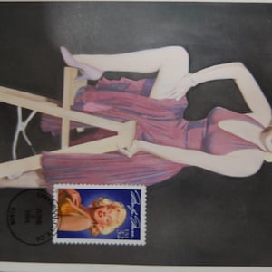 May include: A vintage postcard featuring a stylized image of a woman in a maroon dress, reclining on a wooden structure against a dark background. A postage stamp with a portrait of the same woman is affixed to the postcard. The postcard has a black ink stamp.