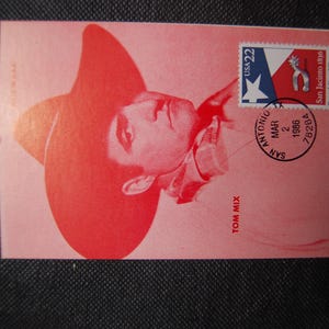 May include: A pink postcard featuring a red image of a person wearing a cowboy hat. The postcard has a USA 22-cent stamp with a Texas flag design and a postmark from San Antonio, Texas, dated March 2, 1986. The name "TOM MIX" is printed on the card.