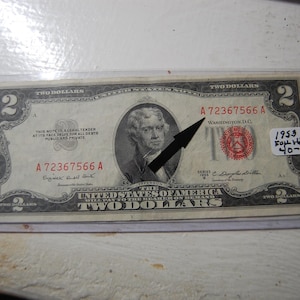 May include: A 1953 two-dollar bill in a protective sleeve. The bill features a portrait of Thomas Jefferson and the text "The United States of America." The bill is in a clear plastic holder.