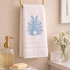 Könnte beinhalten: Weißes Badetuch mit hellblauer Hasen- und Blumenstickerei, an einem goldenen Handtuchring aufgehängt. Ein klarer Seifenspender aus Glas und Seife sind im Badezimmer zu sehen.
