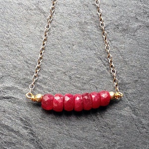 May include: A delicate necklace featuring a strand of faceted, deep red gemstones. The gemstones are strung on a fine gold chain, with small gold beads at each end. The necklace is displayed against a dark gray background.