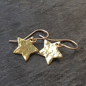 May include: A pair of gold-toned star-shaped earrings featuring a hammered, textured surface. Each star is suspended from a curved gold-toned ear wire hook. The metal has a reflective, polished finish that catches the light across the indentations.