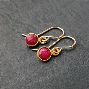 May include: A pair of gold-toned drop earrings featuring round, faceted, deep red stones set in simple metal bezels. The earrings are attached to curved gold-toned ear wires that loop through the top of each circular stone setting.
