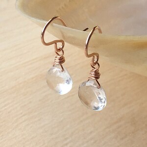 May include: A pair of drop earrings featuring clear, faceted, teardrop-shaped stones suspended from rose gold-toned wire hooks. Each stone is securely attached to the metal hook with a delicate, coiled rose gold wire wrapping at the top.