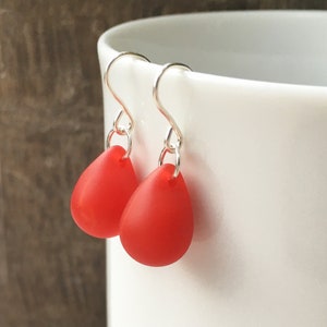 Peut inclure: Une paire de boucles d'oreilles pendantes composées de perles rouges mates en forme de goutte d'eau, suspendues à des crochets en métal argenté. Les boucles présentent un design simple avec une base arrondie et un anneau de jonction.