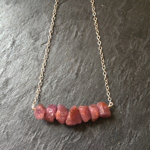 May include: A necklace featuring a delicate silver-toned metal chain connected to a horizontal row of seven raw, unpolished, deep pinkish-red gemstone beads. The stones have an irregular, organic shape and a textured surface, suspended from the chain.