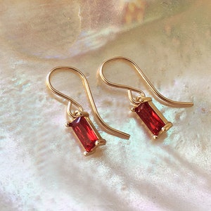 May include: A pair of gold-colored earrings with a hook design. Each earring features a rectangular, vibrant red gemstone set in a gold frame. The earrings are displayed against a pearlescent, off-white background.