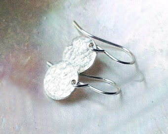 May include: A pair of silver-toned drop earrings featuring small, circular discs with a textured, hammered surface. Each disc is attached to a curved ear wire, creating a simple dangle design that reflects light across the uneven metal finish.