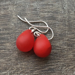 May include: A pair of red, teardrop-shaped earrings with silver hooks. The earrings have a matte finish and are displayed on a wooden surface. These earrings are a simple, elegant accessory.