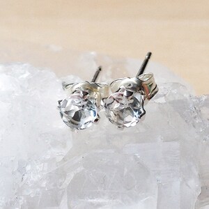 May include: A pair of silver stud earrings featuring clear, faceted gemstones. The earrings are displayed on a white, crystalline surface, creating a sparkling effect. The studs are small and delicate, suitable for everyday wear.