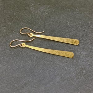 May include: A pair of gold-colored, hammered metal earrings. Each earring features a curved hook and a long, slender, rectangular pendant with rounded edges. The earrings are displayed on a dark gray surface.