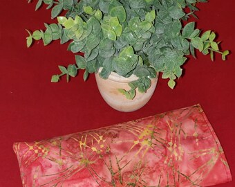 May include: A rectangular fabric eye pillow featuring a red and pink batik-style pattern with thin, intersecting yellow lines. The pillow is positioned below a small, potted artificial green plant with rounded leaves in a light-colored pot.