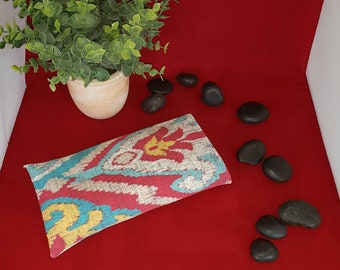 May include: A rectangular fabric pouch featuring a vibrant ikat pattern in shades of red, turquoise, and yellow. The textile displays a repeating abstract design with soft, blurred edges, characteristic of traditional ikat weaving techniques.