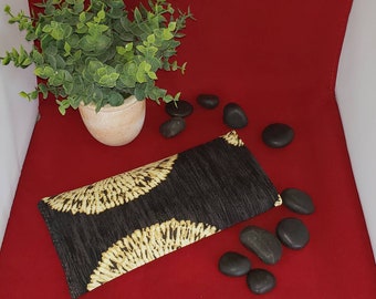 May include: A rectangular black clutch featuring a textured fabric surface with two prominent, semi-circular gold-colored patterns. The design displays a repeating, radiating line motif in a light gold hue against the dark, fibrous background.