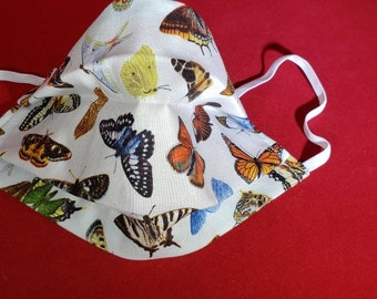 May include: A white fabric face mask featuring a colorful all-over print of various butterfly species in shades of orange, yellow, blue, and black. The mask has a structured center seam and includes two white elastic ear loops on the sides.