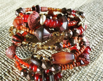 May include: A multi-strand wrap bracelet featuring a variety of beads in shades of red, brown, and bronze. The design includes faceted, smooth, and textured beads, along with a small metallic starfish charm and a hammered silver-tone disc.