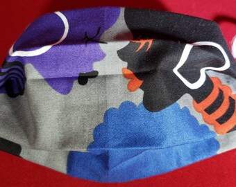 May include: A pleated fabric face mask featuring a graphic print of abstract faces in purple, black, blue, and orange on a gray background. The mask includes white elastic ear loops on both sides for secure placement over the nose and mouth.