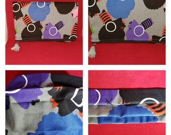 May include: A rectangular fabric clutch bag featuring a repeating pattern of stylized faces in black, blue, and purple on a gray background. The bag includes a dark zipper closure with a light gray tassel pull attached to the side for easy handling.