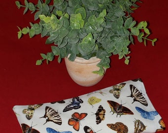 May include: A rectangular white fabric pillow featuring a detailed print of various butterflies in shades of orange, blue, black, yellow, and brown. The pillow is positioned below a small potted green plant with rounded leaves.