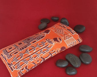 May include: A rectangular fabric pouch featuring a vibrant orange background with a dark brown repeating pattern of stylized figures and geometric shapes, surrounded by several smooth, dark gray river stones on a solid red surface.