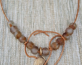 May include: A necklace featuring a thin copper-colored wire cord with a gold-toned metal clasp. The cord is adorned with ten translucent, frosted brown beads and a central oval-shaped gold-toned metal pendant depicting a stylized face.