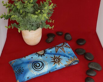 May include: A rectangular fabric eye pillow featuring a blue tie-dye background with gold and black sun, star, and spiral motifs. The pillow has a smooth, soft texture and is designed with a repeating celestial pattern across the entire surface.