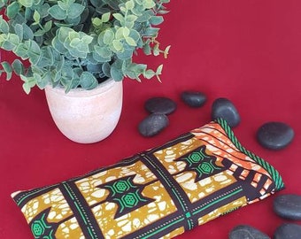 May include: A rectangular fabric pouch featuring a repeating geometric pattern in mustard yellow, black, and white, accented with green and orange stripes. The pouch is positioned near a small potted green plant and several smooth black stones.