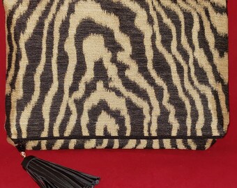 May include: A rectangular clutch bag featuring a textured black and gold zebra-like pattern. The bag has a fold-over design and includes a black faux leather tassel attached to the zipper pull, which is finished with a small metallic gold bead.