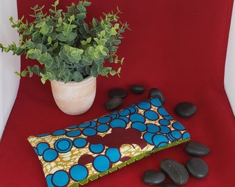May include: A rectangular fabric pouch featuring a vibrant pattern of blue circles outlined in dark brown on a cream background, with a green and yellow striped edge detail. The pouch is positioned horizontally on a deep red surface.