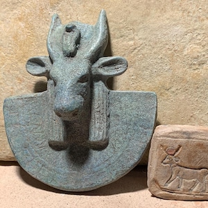 Könnte beinhalten: Eine grüne, texturierte, halbkreisförmige Wandplatte mit einem skulptierten Stierkopf mit markanten Hörnern und Ohren, neben einer kleinen, rechteckigen, beigefarbenen Steinplatte mit einer eingravierten Abbildung eines Stiers mit Sonnenscheibe.