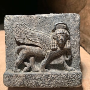 Puede incluir: Un relieve de piedra tallada que representa una esfinge, criatura mitológica con cabeza humana y cuerpo de león, con grandes alas. La escultura gris presenta detalles intrincados, mostrando arte antiguo.