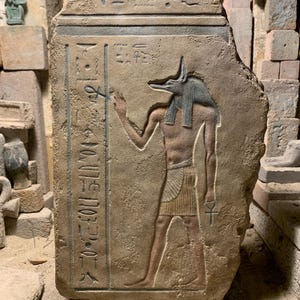 May include: A rectangular stone relief carving featuring a standing jackal-headed figure in profile, holding an ankh symbol. The figure is surrounded by vertical columns of incised Egyptian hieroglyphs, with a textured, weathered tan surface.